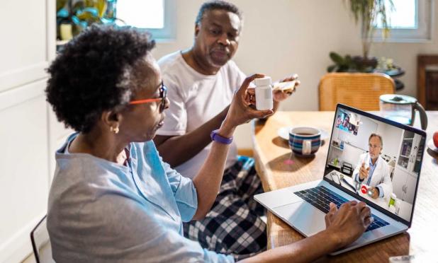 Person on laptop at kitchen table with another who holds up prescription bottle for telehealth doctor 