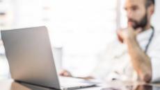 Doctor at desk sits back and reads information on a laptop